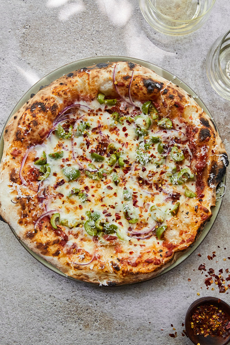 A full shot of a pizza topped with green olives and a bit of crushed red pepper in the corner.