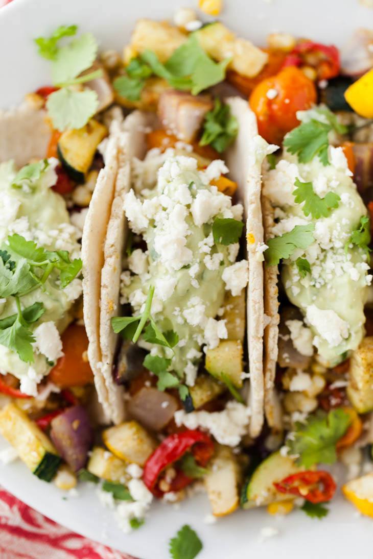 Close-up, overhead photo of tacos on a white plate, loaded with roasted summer vegetables, an avocado cream, and a sprinkle of feta.