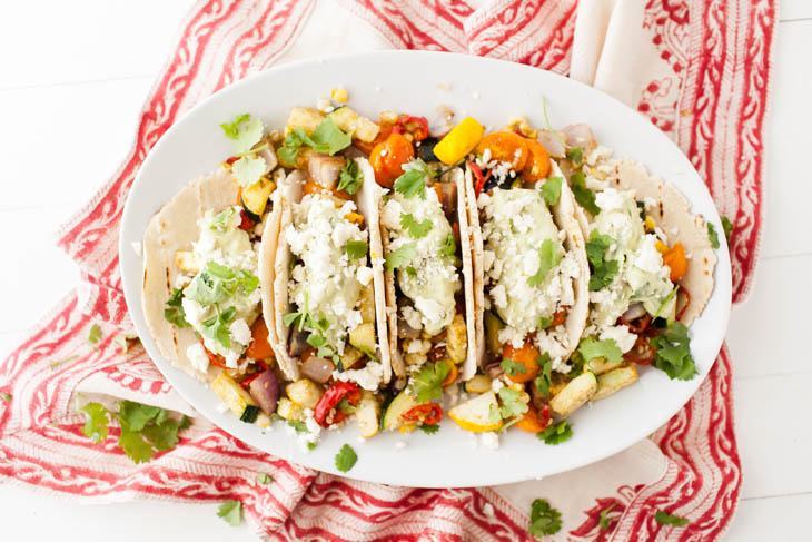 Overhead shot of tacos on a white plate, loaded with roasted summer vegetables, an avocado cream, and a sprinkle of feta.