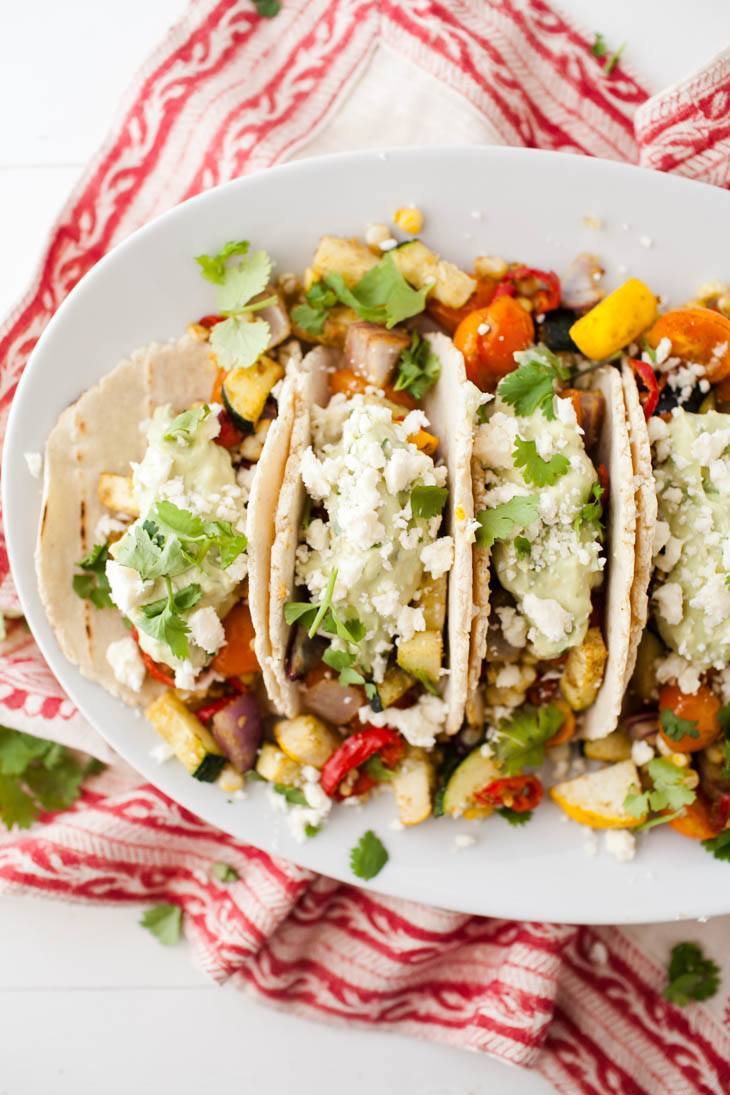 Overhead shot of tacos on a white plate, loaded with roasted summer vegetables, an avocado cream, and a sprinkle of feta.