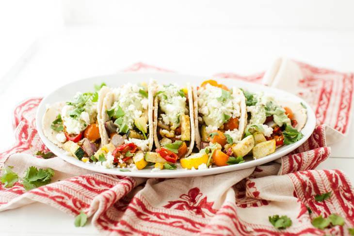 Side-angle photo of tacos on a white plate, loaded with roasted summer vegetables, an avocado cream, and a sprinkle of feta.