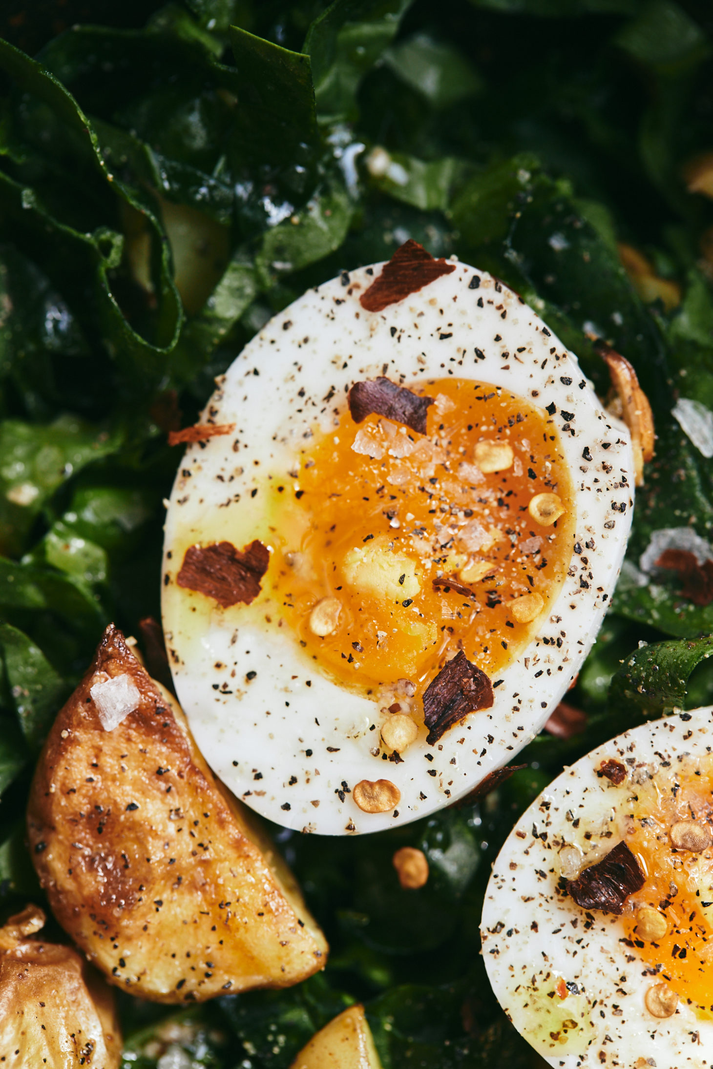 Close-up Overhead photograph of a spinach salad topped with roasted potato wedges and soft-boiled eggs.