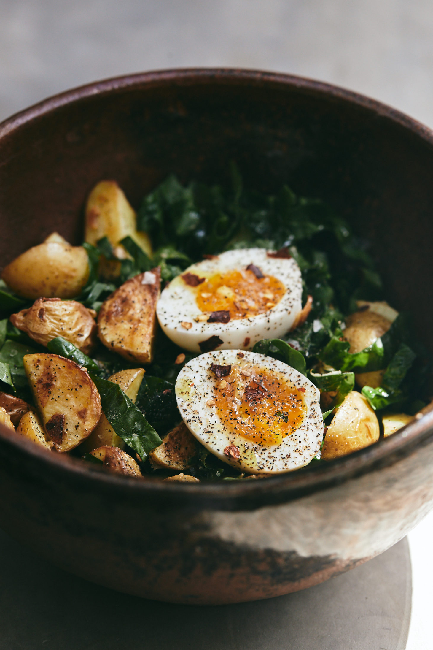 Side-angle photograph of a spinach salad topped with roasted potato wedges and soft-boiled eggs.