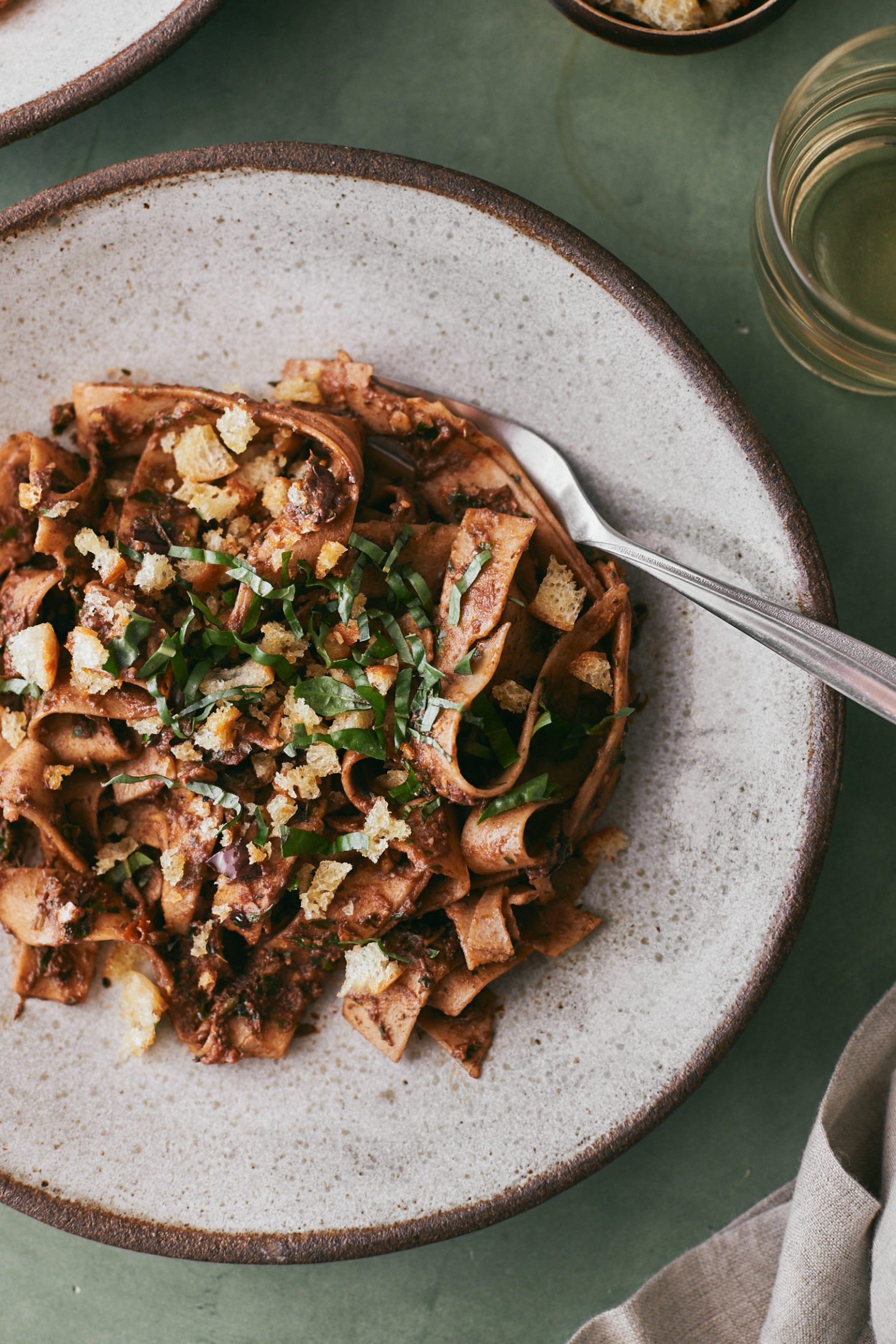 Close-up photo of a grey-speckled bowl with pasta, olive pesto, breadcrumbs, and topped with shredded basil.