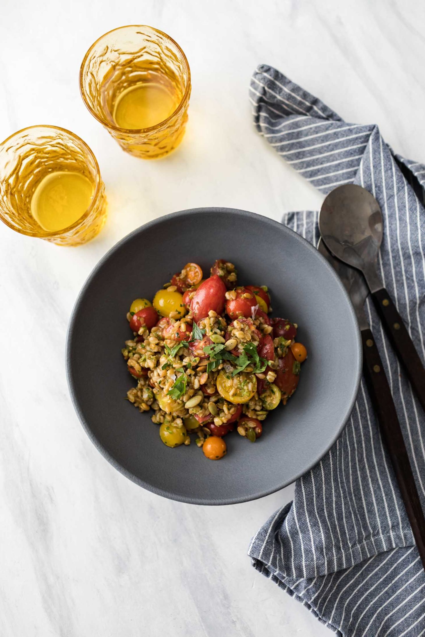 Overhead, far-away shot of two yellow glasses, a stripped blue napkin, and a blue-grey bowl with a grape tomato grain salad with green harrisa.