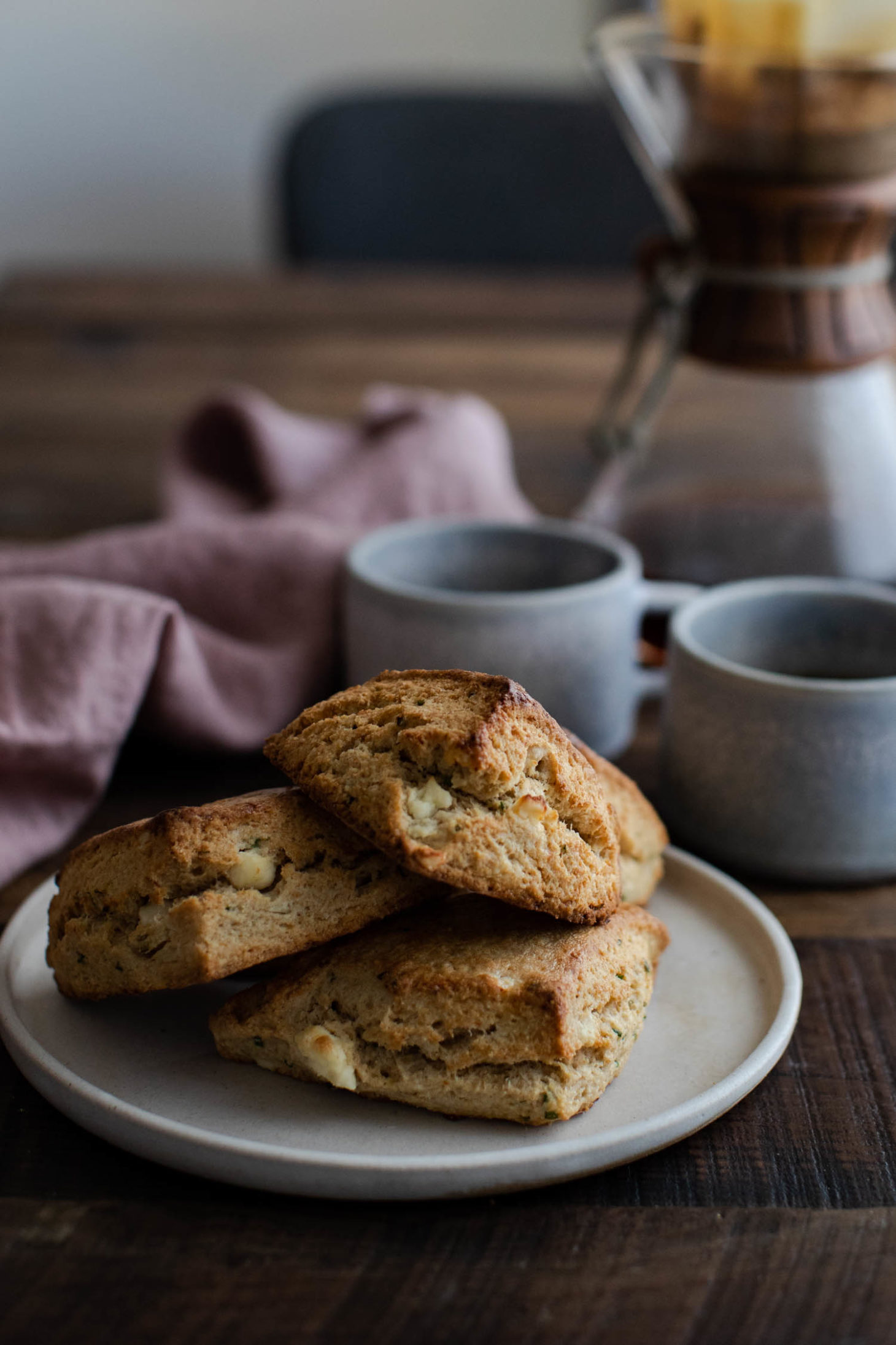 Side angle photo of golden, savory scones filled with feta and chives with blue coffee mugs and chemex in the background.