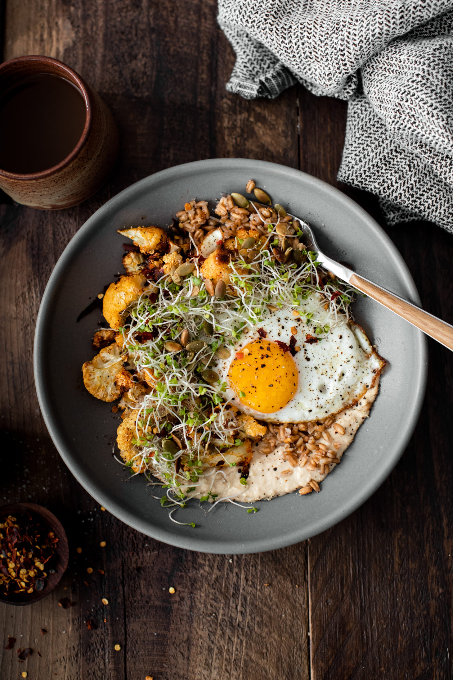 Overhead photograph of a grey bowl with fried egg on-top of paprika roasted cauliflower, farro, and hummus.