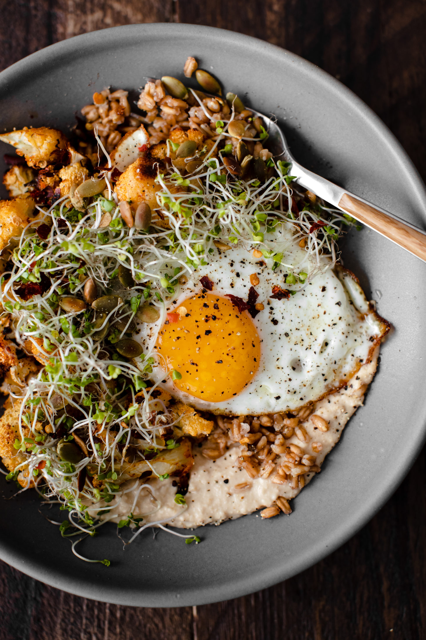 Close-up overhead photograph of a grey bowl with farro, hummus, roasted cauliflower, and a fried egg.
