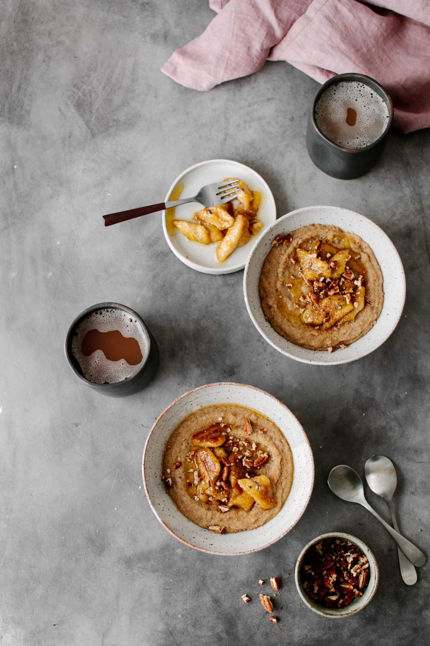 Overhead photograph of amaranth porridge in white bowls topped with caramelized bananas and served with coffee in black mugs.