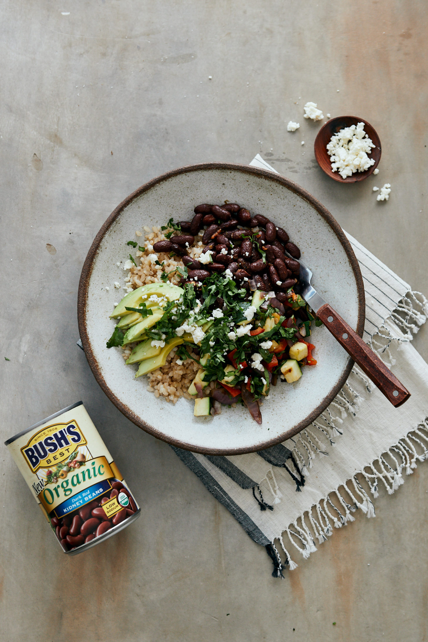 Light grey background with a small bowl of crumbled cheese and a large bowl filled with brown rice, grilled vegetables, kidney beans, and avocado.