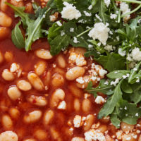 Close-up image of white beans in a tomato broth with arugula and feta on top.