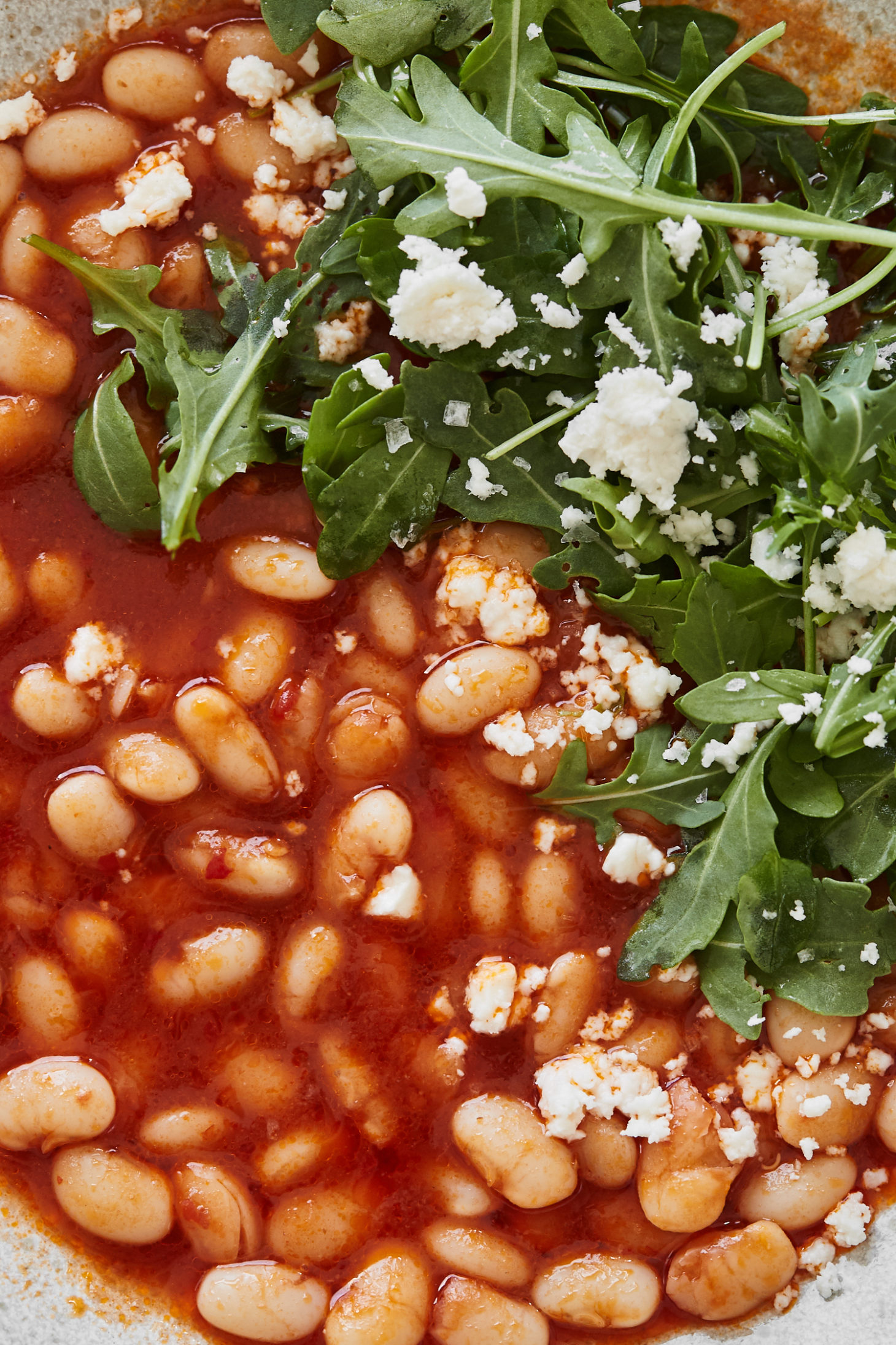 Close-up image of white beans in a tomato broth with arugula and feta on top.