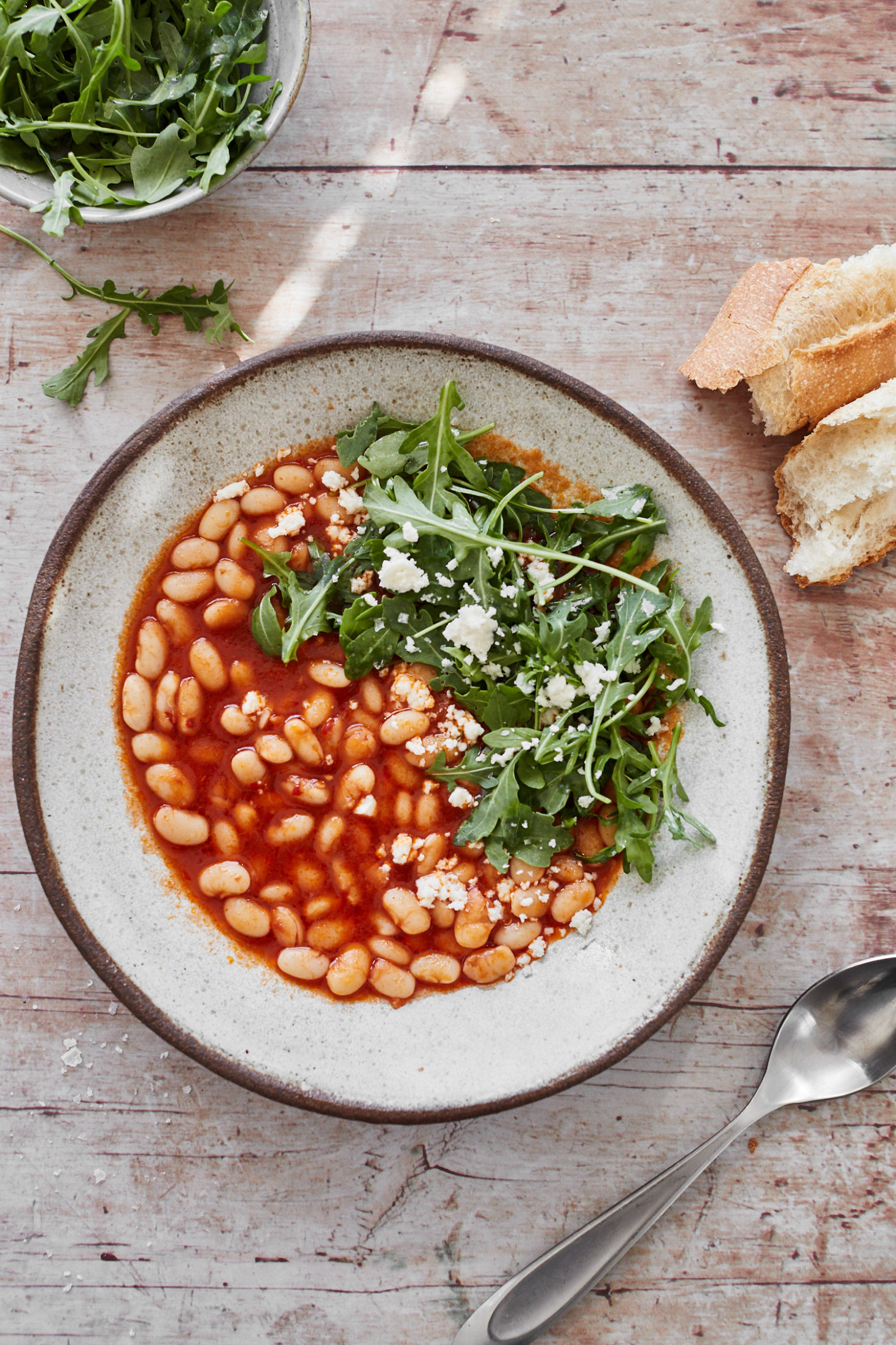 Light image with a bowl of tomato bean stew with arugula on top on a white background.