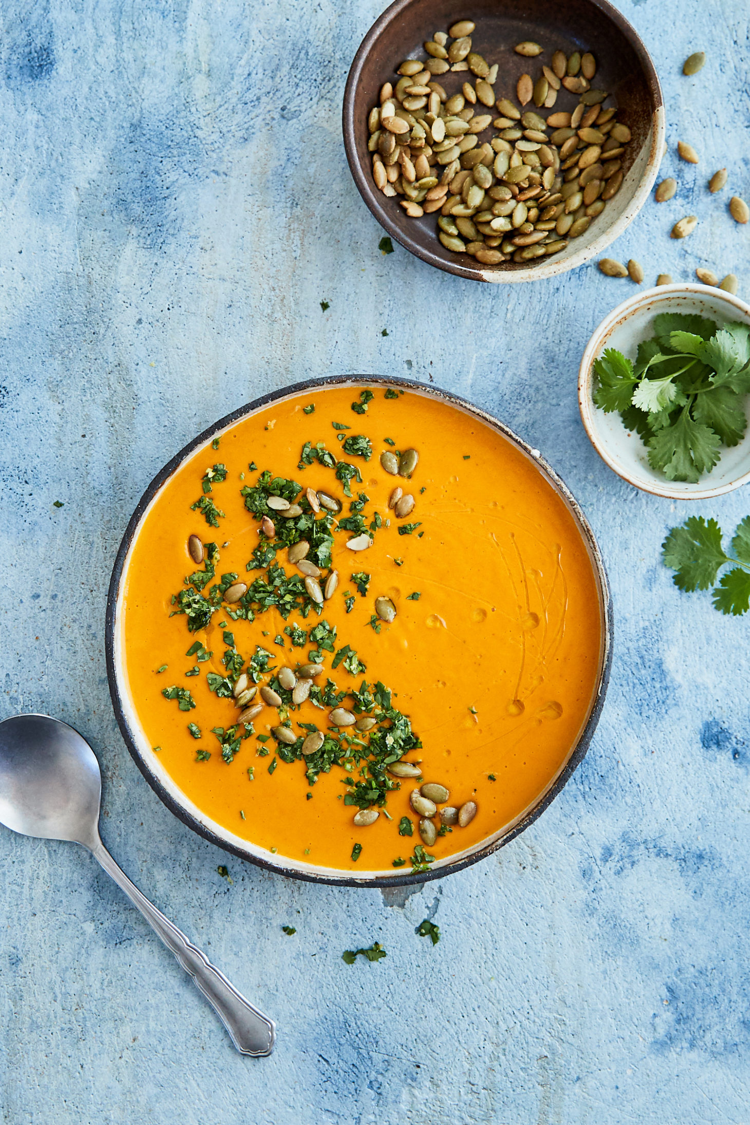 Photo of carrot soup on a blue background with small bowls of pepitas and cilantro around the bow.