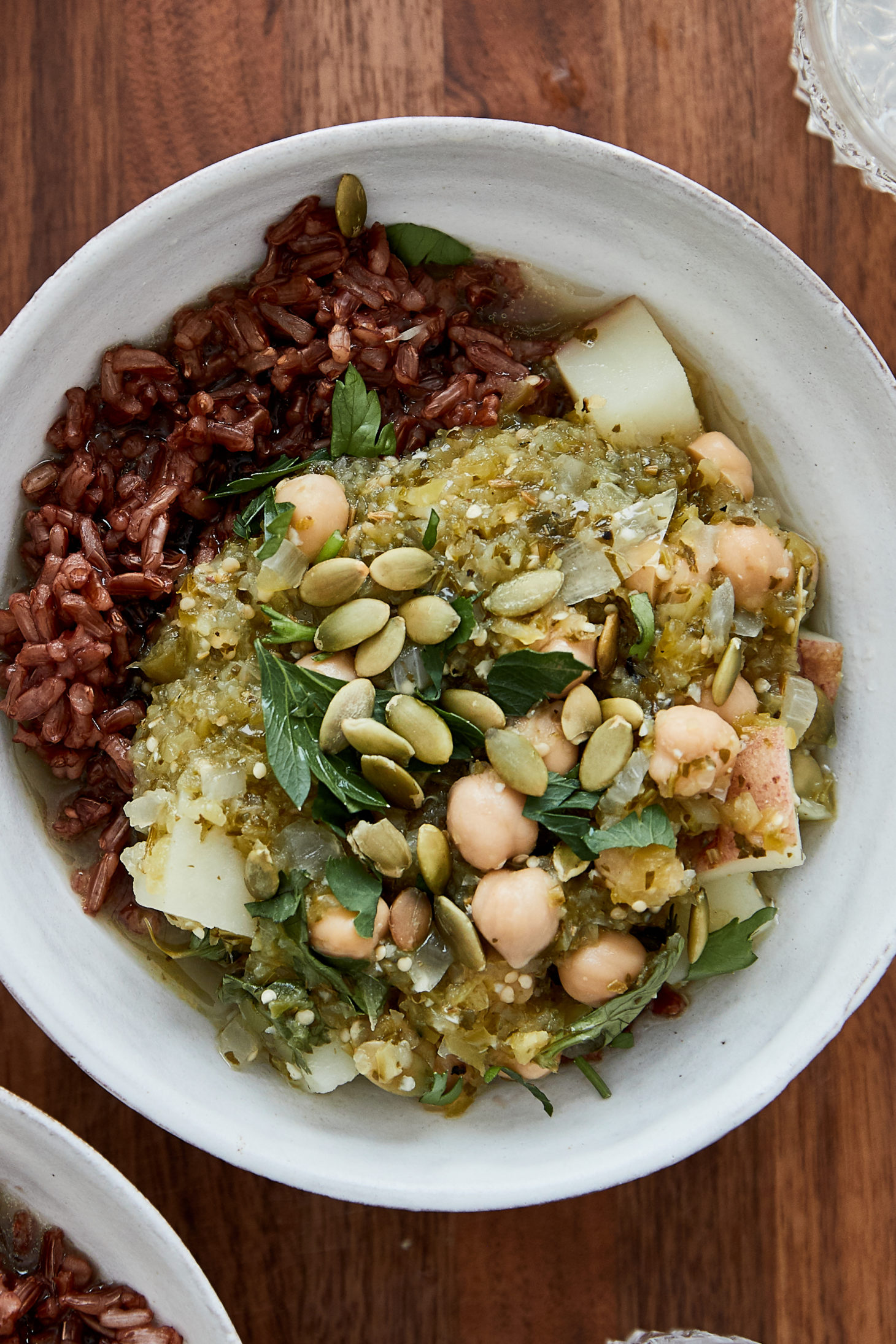 Close-up photo of a white bowl filled with red rice and a green vegetarian chickpea chili on a wood background.