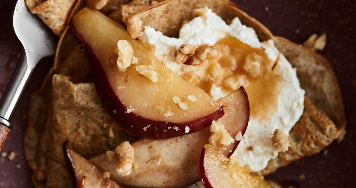 Close-up of rye crepes on a maroon plate topped with pears, whipped crepe, and walnuts.