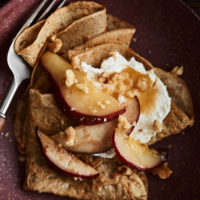 Close-up of rye crepes on a maroon plate topped with pears, whipped crepe, and walnuts.