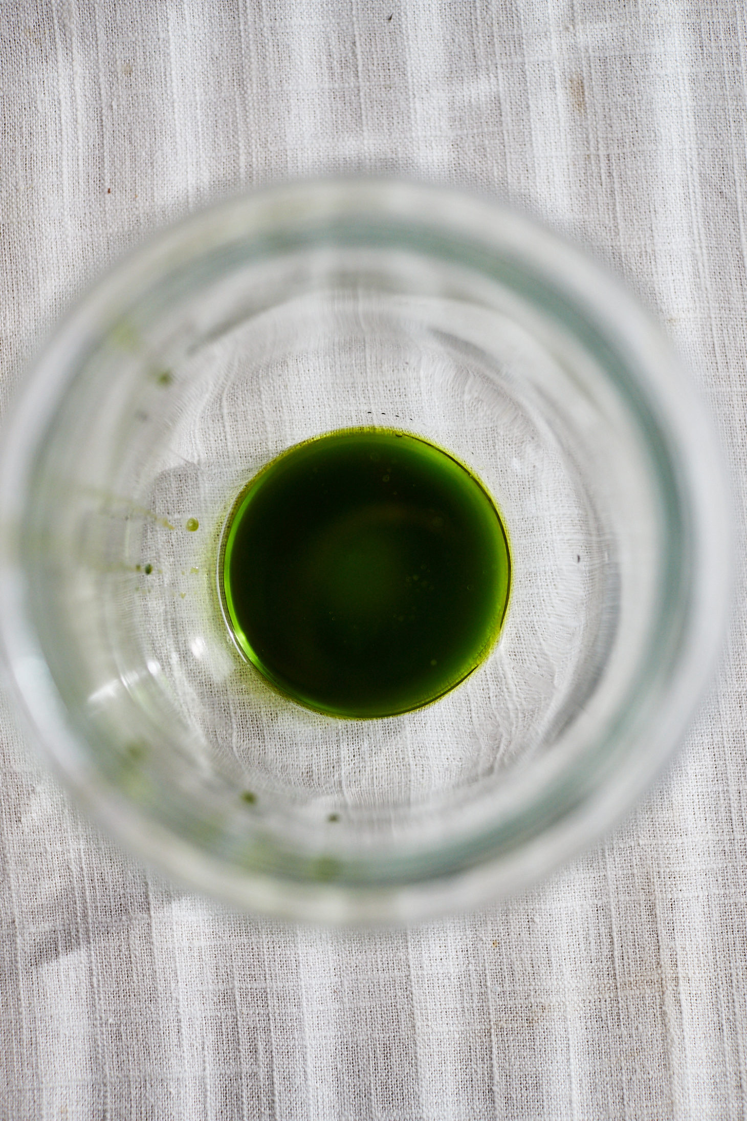 Top-down image of a clear jar on a white linen surface with green parsley oil.