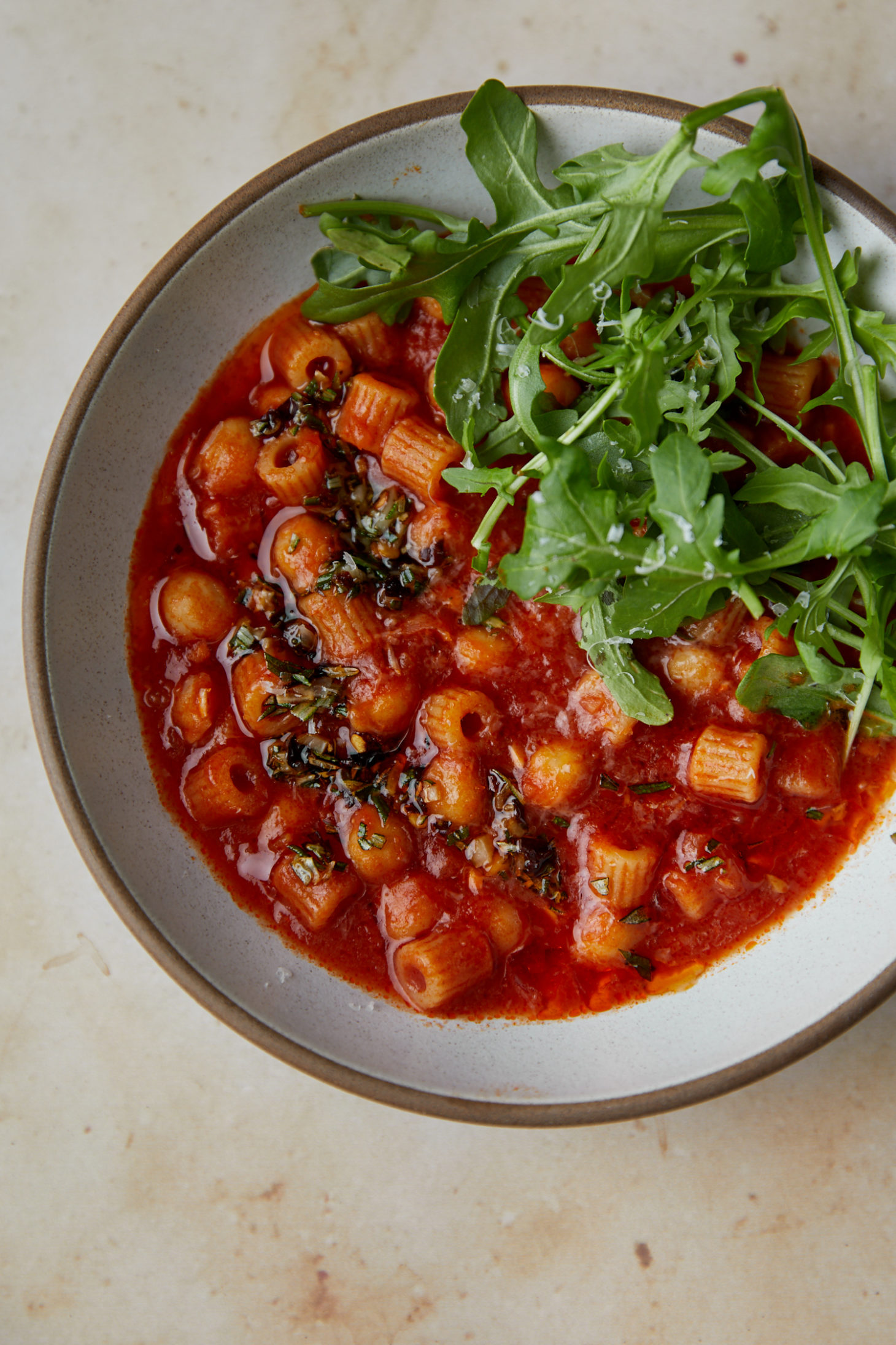 Close-up photo of a greyish bowl filled with pasta, chickpea, and tomato soup with arugula on top.