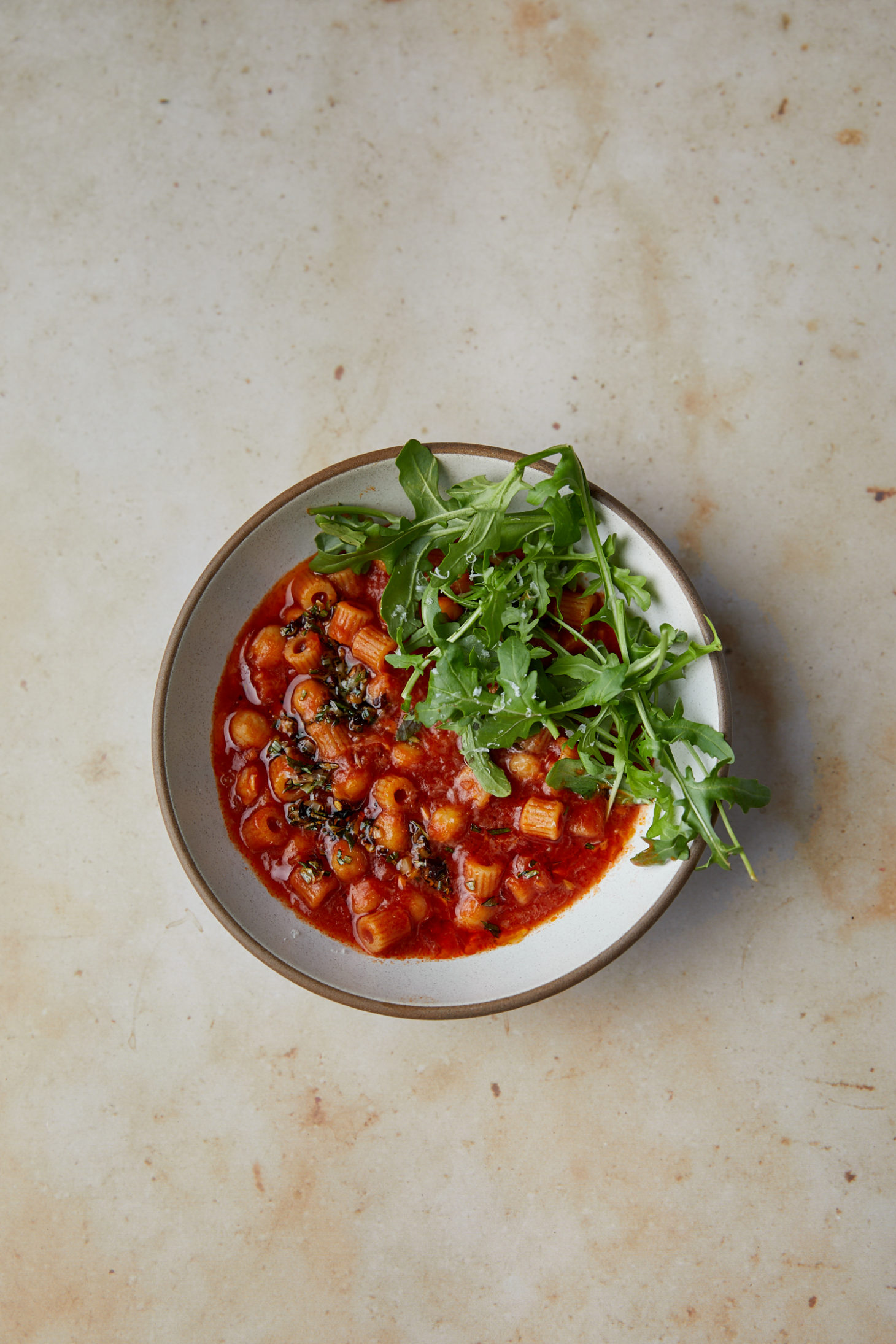 Yellowish marble background with a grey bowl filled with tomato, pasta, and chickpeas.