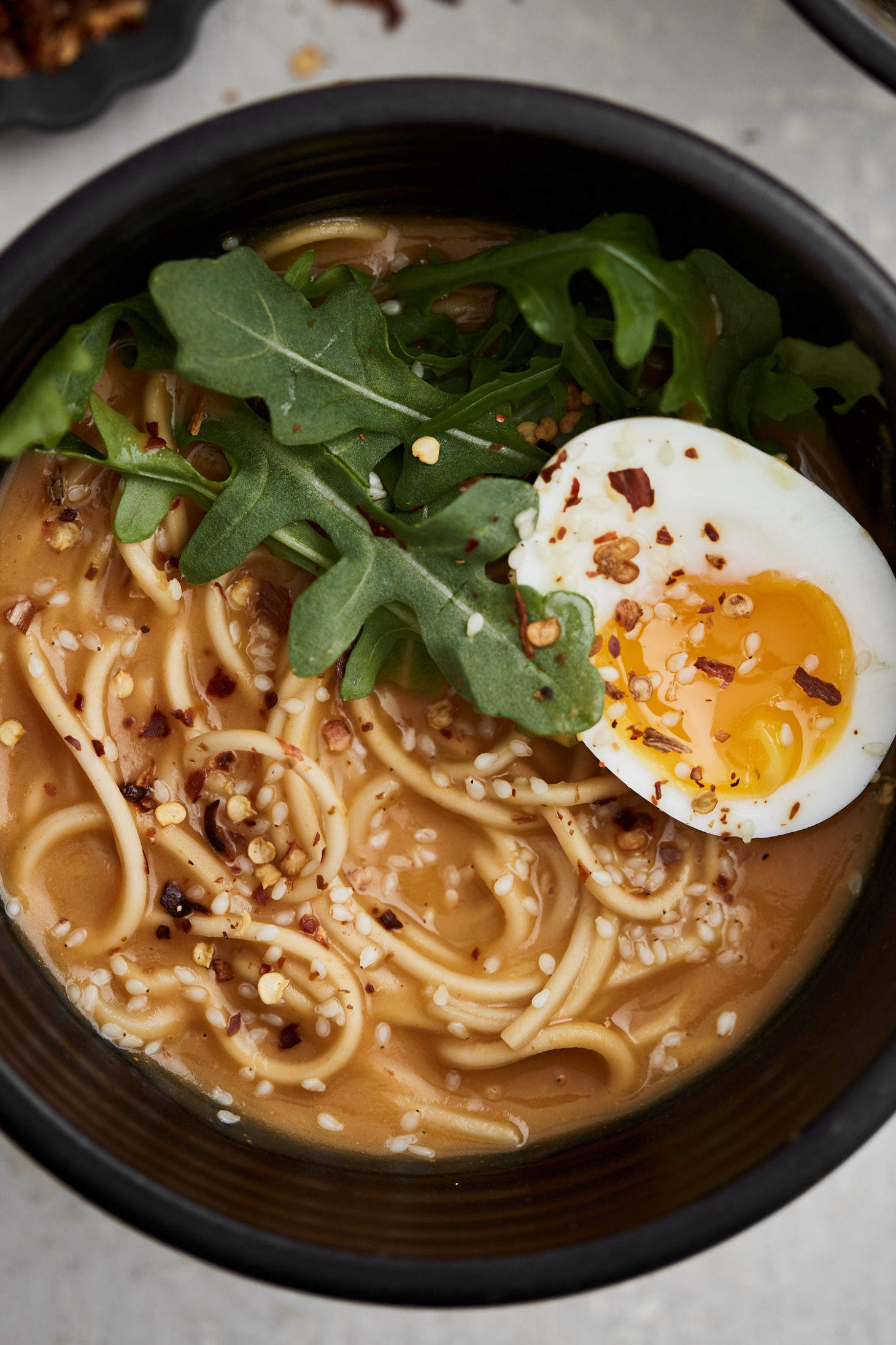 Black bowl filled with noodles, orange broth, arugula, and an egg sliced in half on a light grey background