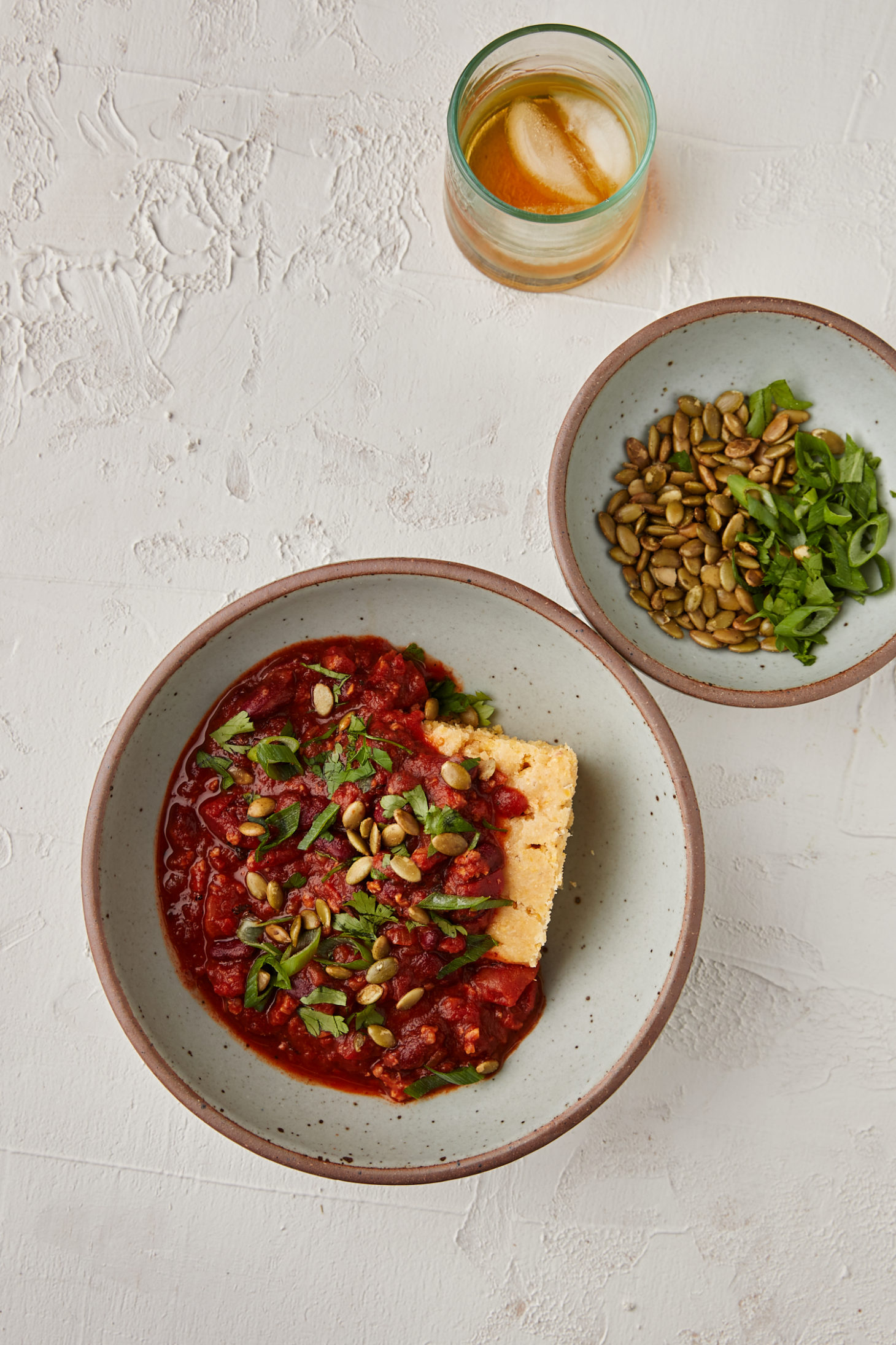 Vegan chili in a grey bowl with a small grey bowl filled with cilantro and pumpkin seeds on a white background.