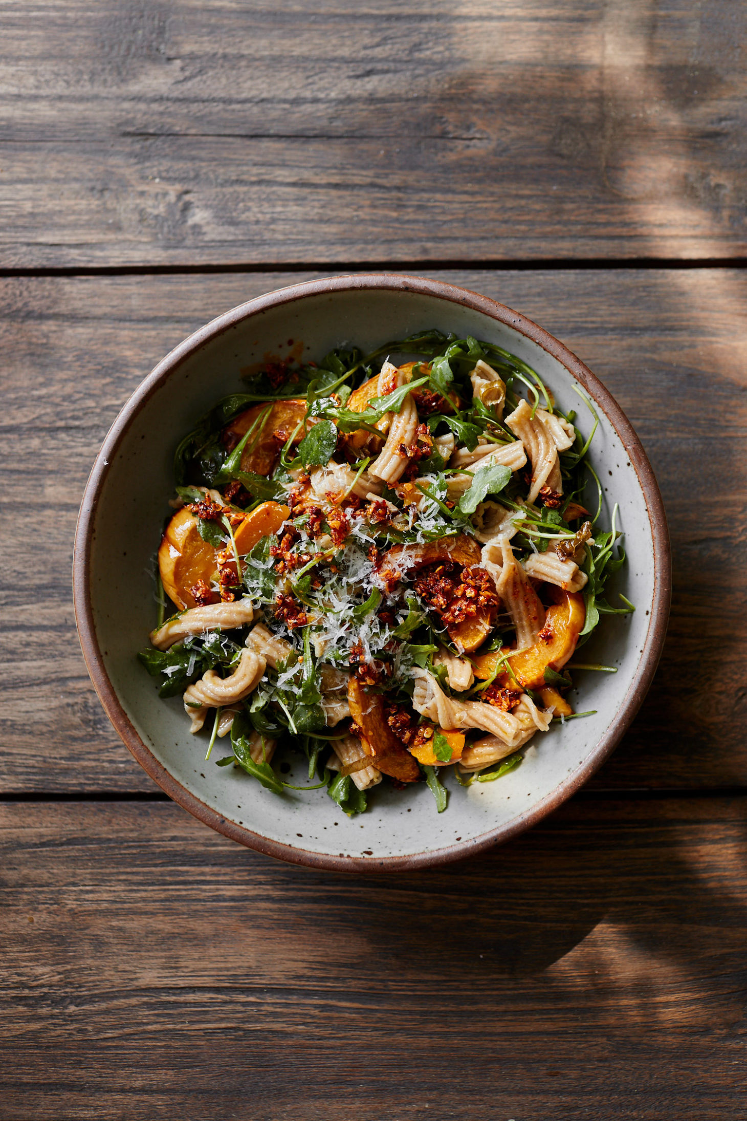 Image of a grey bowl on a light wood table filled with arugula, delicata squash, and pasta.