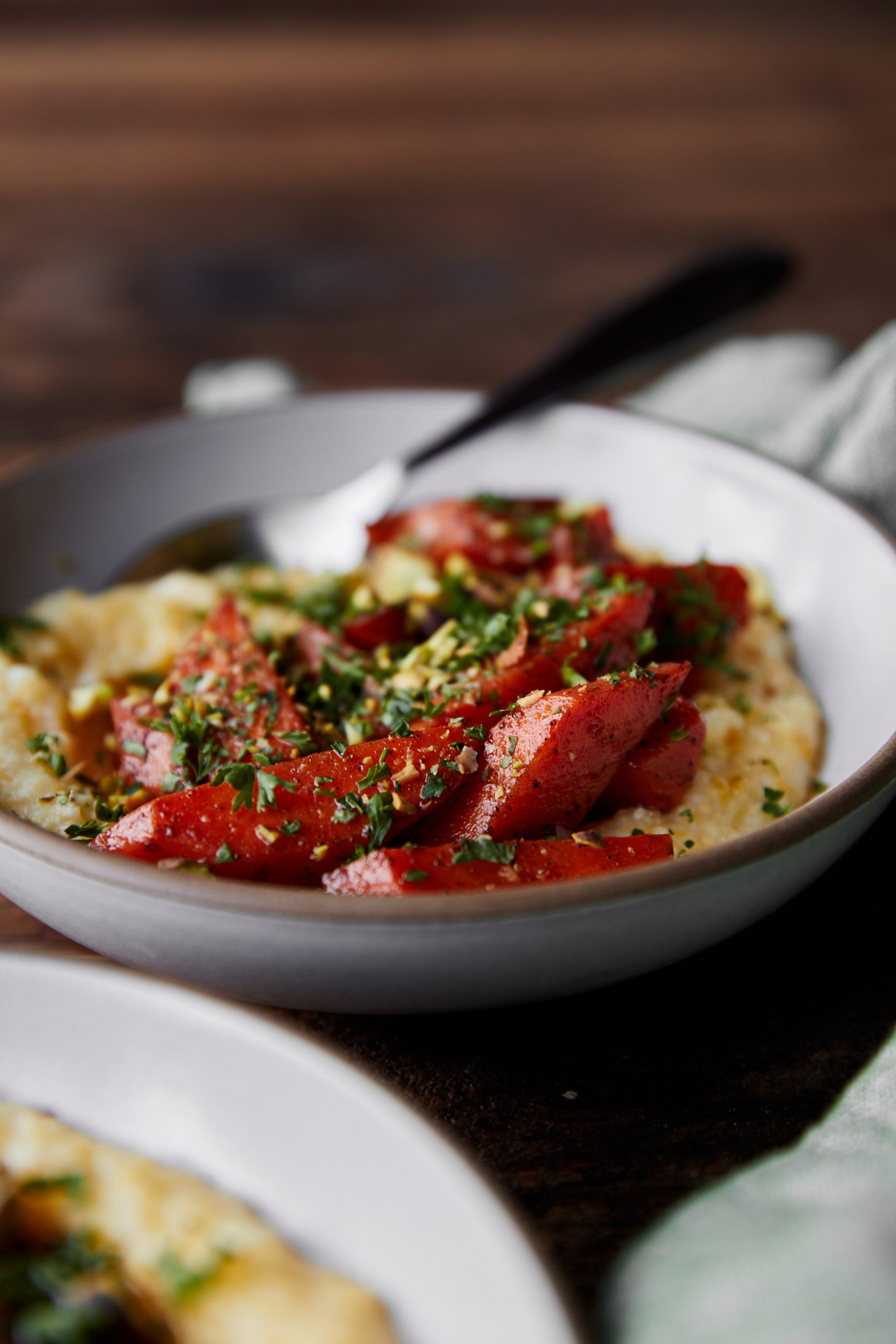 Side photograph of white bowl with yellow polenta topped with orange braised carrots and parsley.