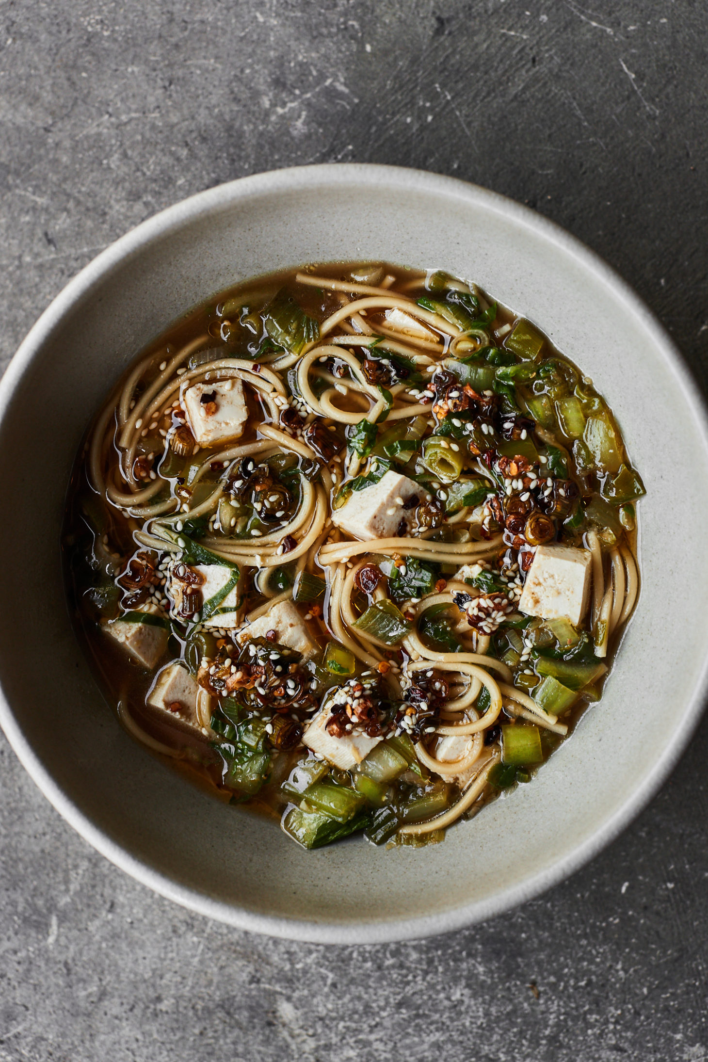A grey bowl on a grey background with noodles, broth, scallions, and chili oil