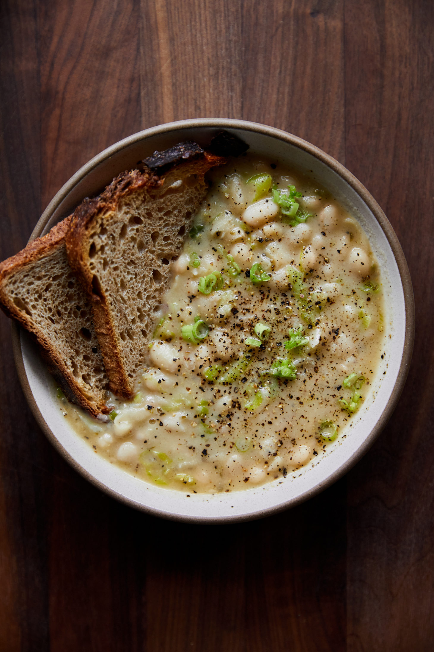 A white bowl on a dark wood counter filled with white beans in a cream-colored broth and toast tucked into the side.