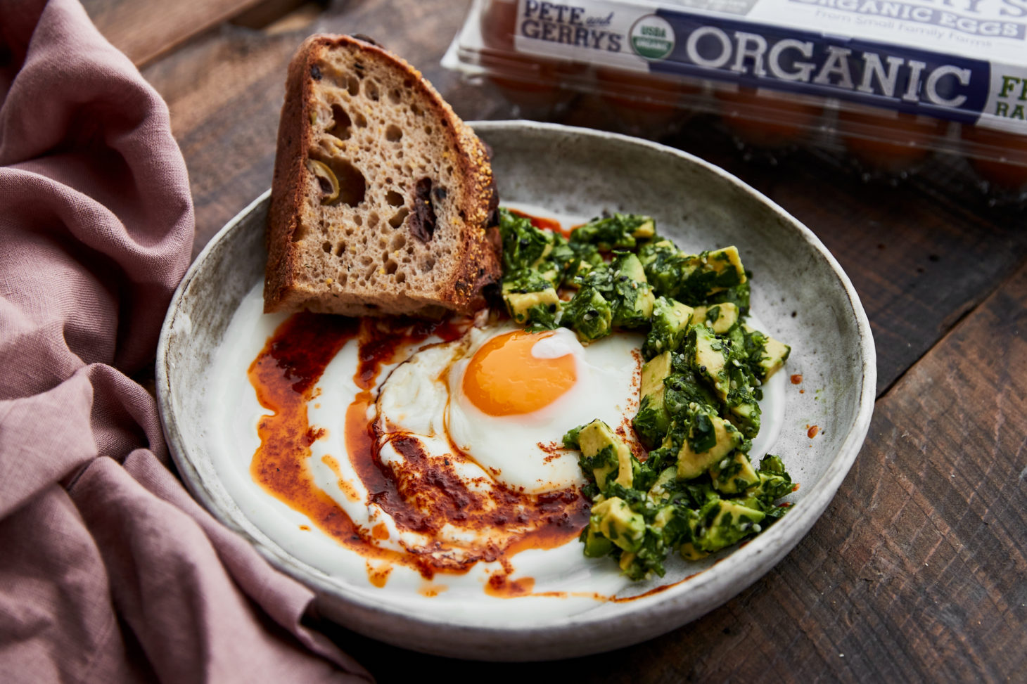 Side-angle photograph of a greyish bowl that has yogurt topped with a sunny-side up egg, avocado relish, and a fiery red infused butter.