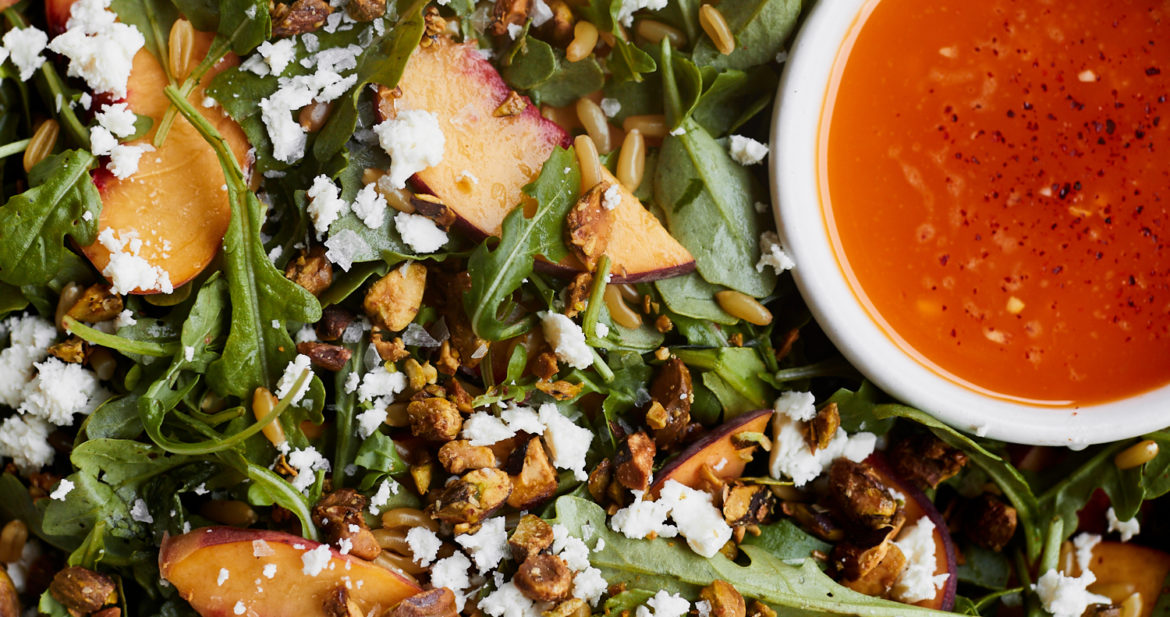 Close-up photo of a salad with peaches, pistachios, a chili dressing- all in a grey bowl.