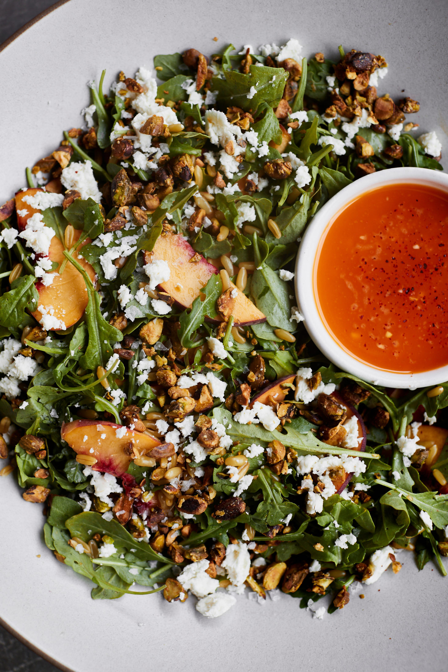 Close-up photo of a salad with peaches, pistachios, a chili dressing- all in a grey bowl.