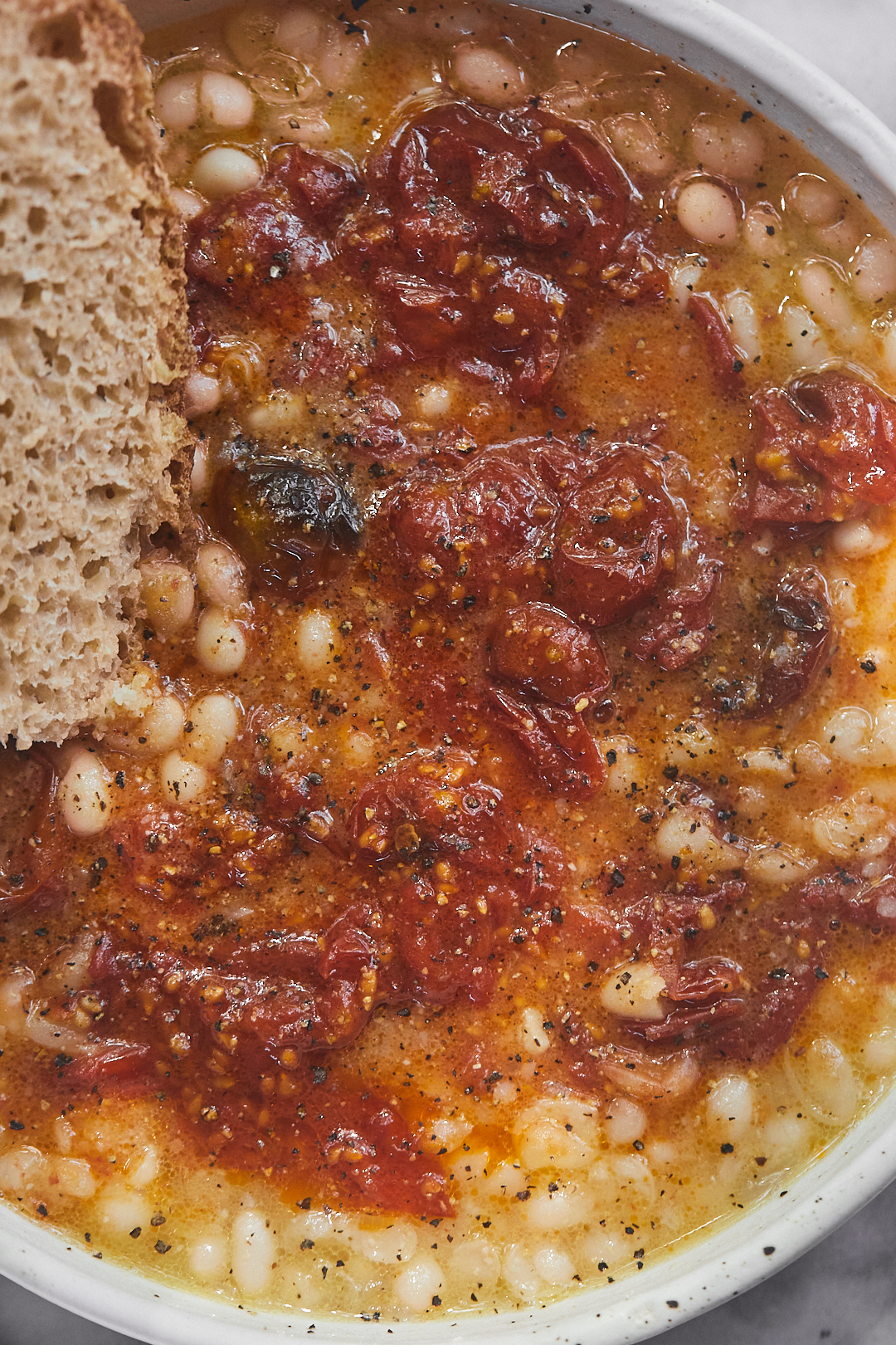 A close-up image of brothy beans accompanied by roasted tomatoes and a slice of bread. 