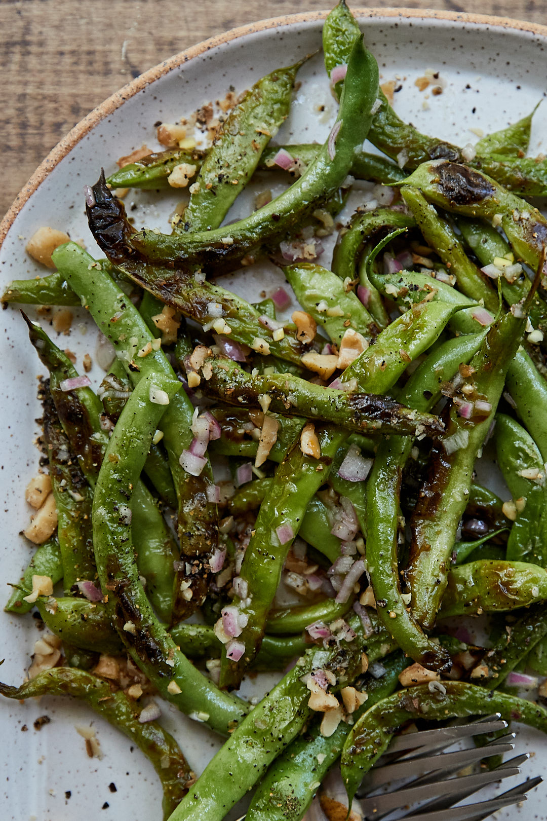 Close-up of charred green beans dotted with walnuts and shallots on a speckled white plate.