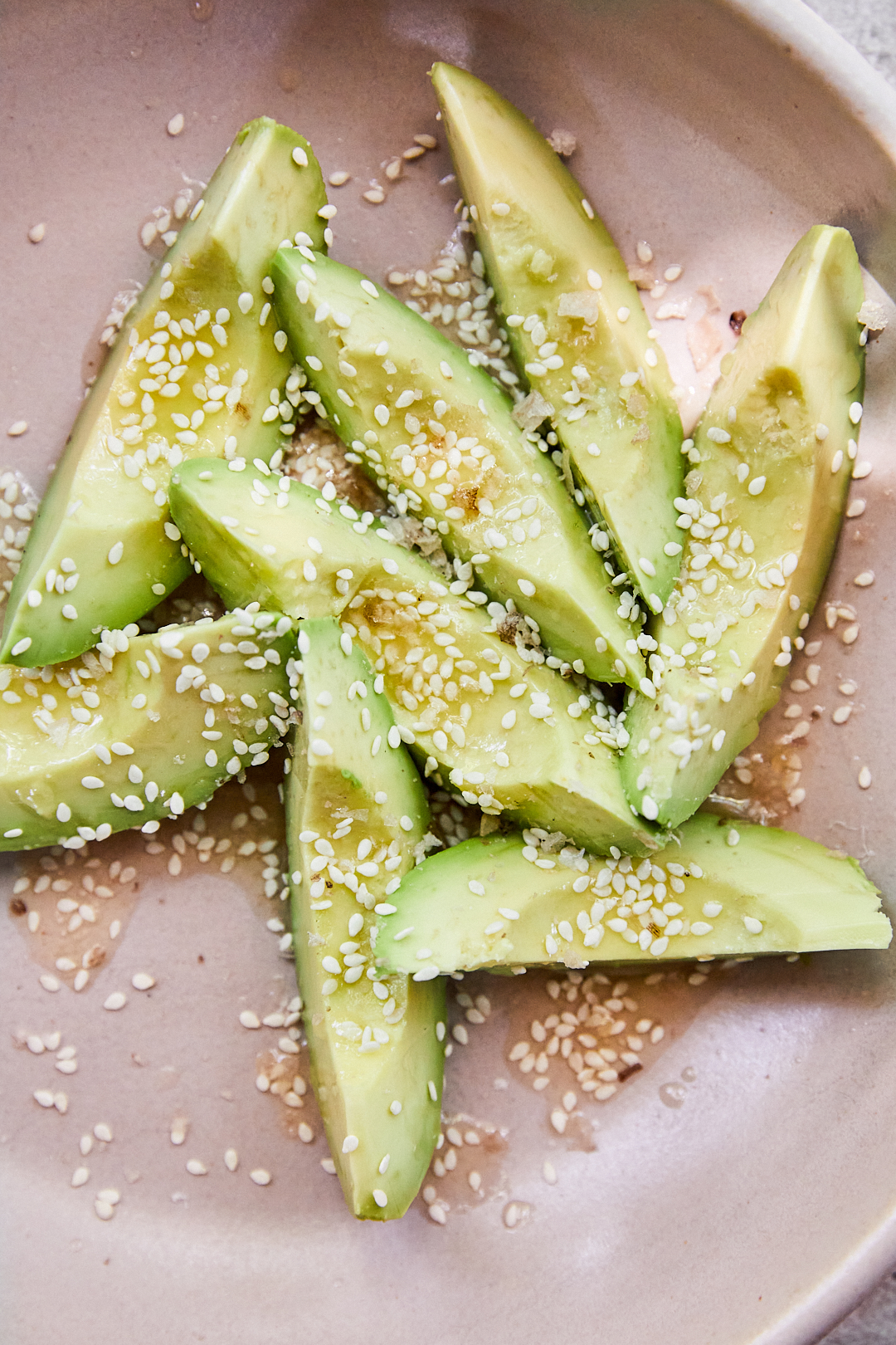 A blush bowl containing wedges of avocado that are topped with sesame seeds, vinegar butter, and pieces of finishing salt. 