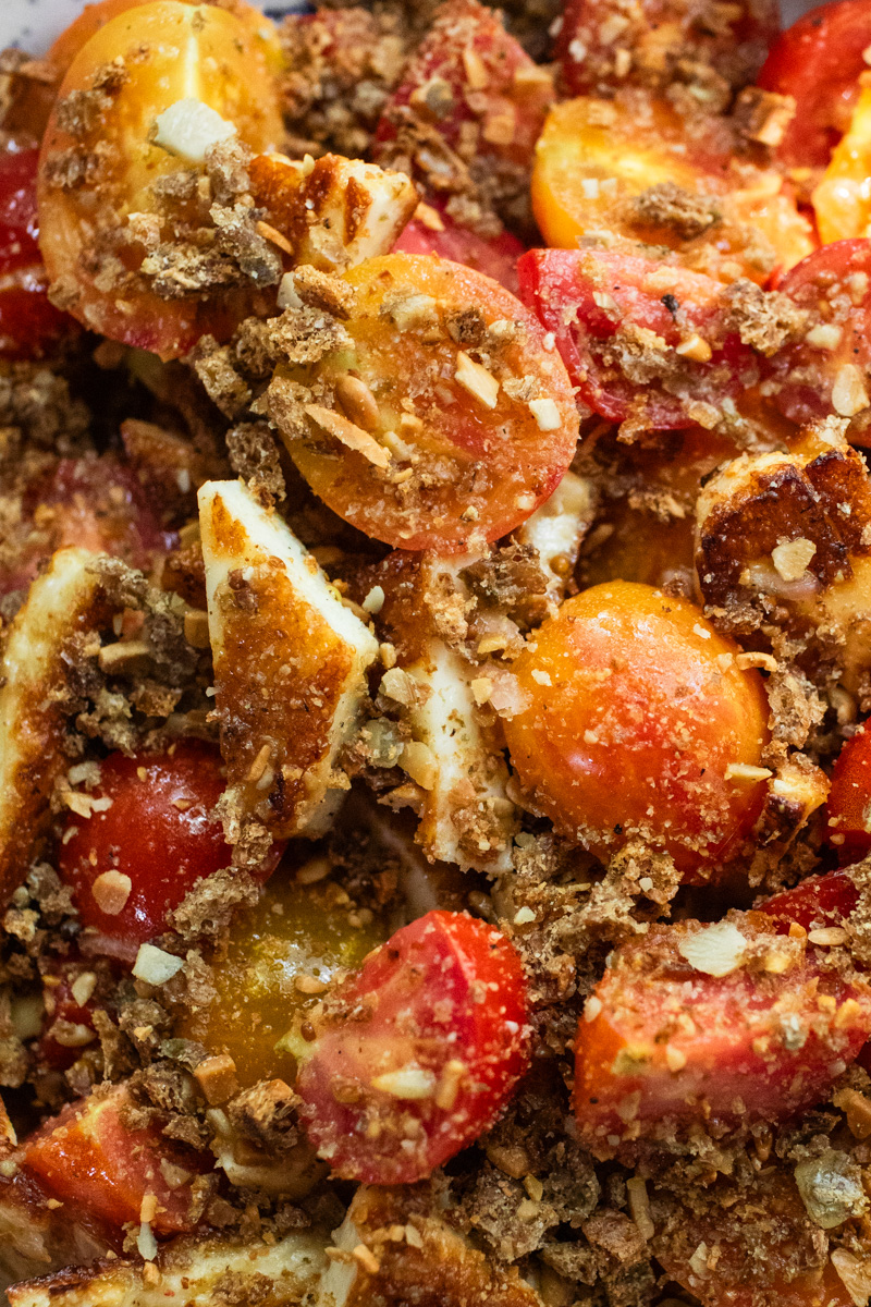 A close-up photo of a halloumi salad featuring sliced red and orange cherry tomatoes and crushed rye crackers.